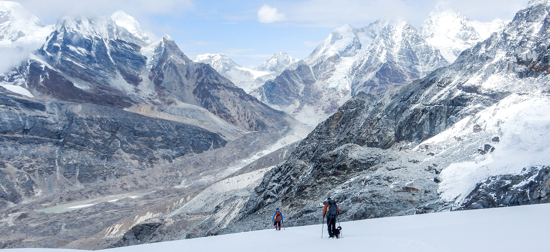 Two trekkers and a dog walking on snow on the way to Mera Peak Base Camp from High Camp with the Hinku Valley in the background.